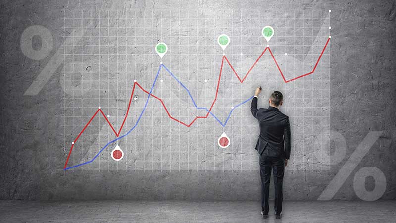 Homem de costas com fato de executivo traça uma linha azul de gráfico num quadro onde já existem outras linhas e símbolos